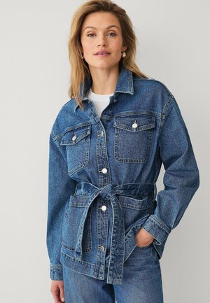 Kobieta ubrana w kurtkę dżinsową w średnim odcieniu zewnętrznym, z przednimi kieszeniami, zapięciem na guziki i przewiązanym paskiem, zestawioną z dopasowanymi dżinsami.