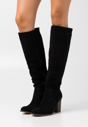 Botas altas de ante negro hasta la rodilla con un tacón de madera grueso, que presentan cremalleras laterales y una silueta ajustada. La textura es suave y lisa.