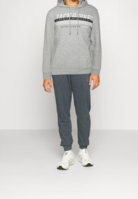 Felpa grigia con stampa "JACK & JONES ATHLEISURE 1990", abbinata a pantaloni tuta grigio scuro e sneakers bianche, dotata di cappuccio con coulisse.