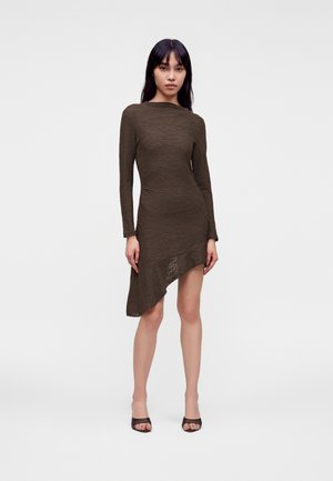 Femme portant une robe marron à manches longues avec un ourlet asymétrique, debout droite devant un fond clair uni.