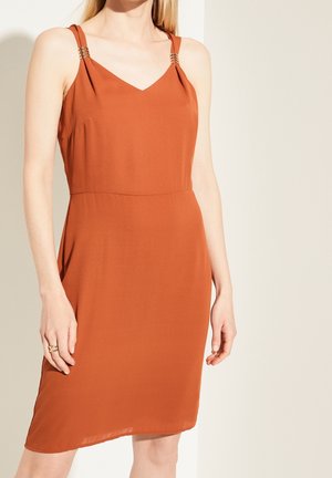 Vrouw die een mouwloze roestkleurige jurk draagt met metalen ringdetails op de schouderbanden, staand tegen een effen beige achtergrond.