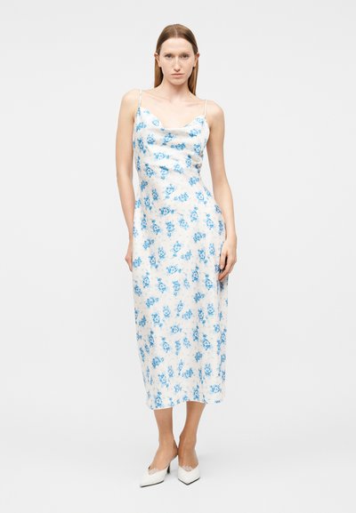 Γυναίκα που φοράει λευκό φόρεμα φόρεμα με μπλε λουλούδια, λεπτές τιράντες και λευκά μυτερά τακούνια, σε απλό φόντο.