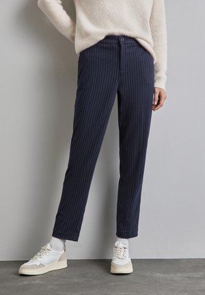 Persoon met marineblauwe pantalon met krijtstrepen, witte sokken, witte sneakers en een crèmekleurige gebreide trui, staand tegen een grijze muur.