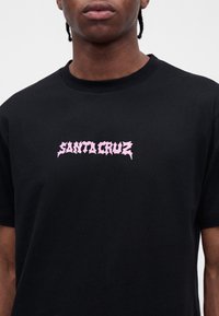 Άτομο που φοράει μαύρο T-shirt με ροζ γοτθικό κείμενο «Santa Cruz» στο στήθος, μπροστά σε απλό φωτεινό φόντο.