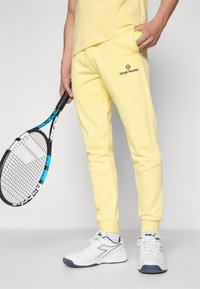 Pantalon de survêtement jaune avec un logo brodé, texture douce, design ajusté et poches. Associé à des chaussures de sport blanches et tenant une raquette de tennis.