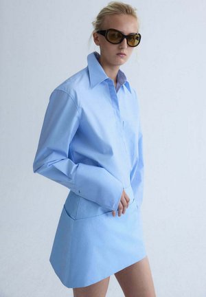 Mujer con vestido tipo camisa azul claro de gran tamaño, con puños largos y gafas de sol con tintado ámbar sobre un fondo claro y liso.