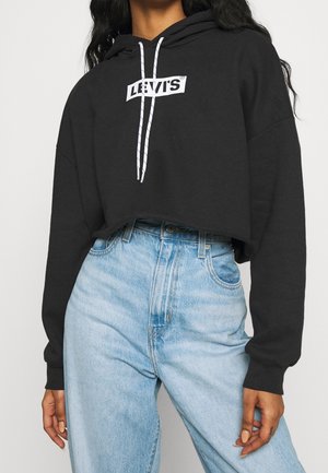 Czarna krótka bluza z kapturem z białym logo Levi's i sznurkami, noszona z jasnoniebieskimi, wysokim stanem jeansami na stojącej osobie.