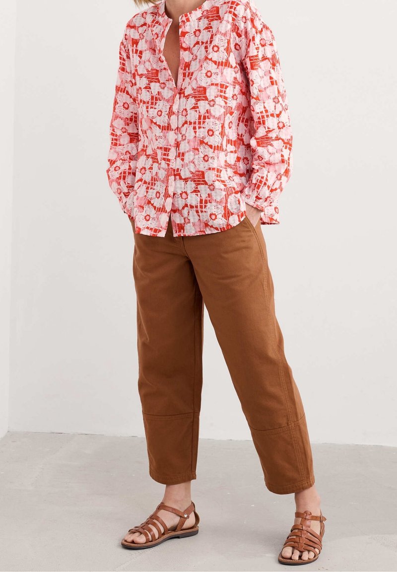 Personne portant un chemisier ample à manches longues rose et rouge à fleurs, un pantalon marron raccourci et des sandales plates marron à brides, debout les mains dans les poches.