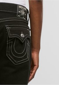 Jeans de mezclilla negros con costuras en contraste blancas, un bolsillo trasero decorativo con un botón y una etiqueta de True Religion en la cintura.