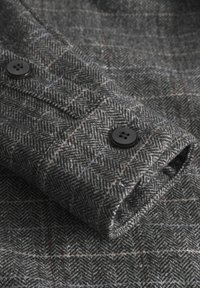 Hallhalline mustriga kangas pehmea tekstuuriga, millel on mustad nupud mansetil ning peened punased ja sinised jooned läbi hõljuvad.