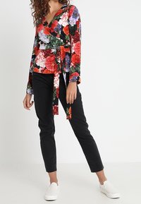 Haut cache-cœur à fleurs avec des fleurs rouges, roses et violettes vibrantes sur un fond noir, manches longues et taille nouée. Porté avec un pantalon noir et des chaussures blanches.