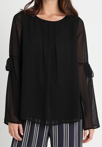 Svart genomskinlig blus med en avslappnad passform, rund halsringning och långa ärmar. Har horisontella veck och rosettaccenter på ärmarnas manschetter.
