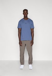 Μπλε βαμβακερό μπλουζάκι με στρογγυλή λαιμόκοψη και μαύρο λογότυπο, συνδυασμένο με μπεζ παντελόνι και λευκά αθλητικά παπούτσια. Το μοντέλο στέκεται μπροστά σε μια απλή πίσω όψη.