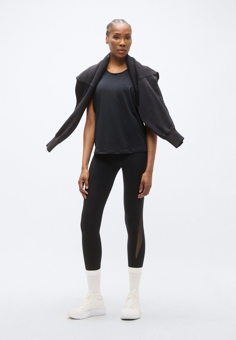 Fekete sportos trikó, fekete leggings átlátszó oldalsó panelekkel és fehér sportcipők. A modell fekete pulóvert visel, amely a vállára van terítve.