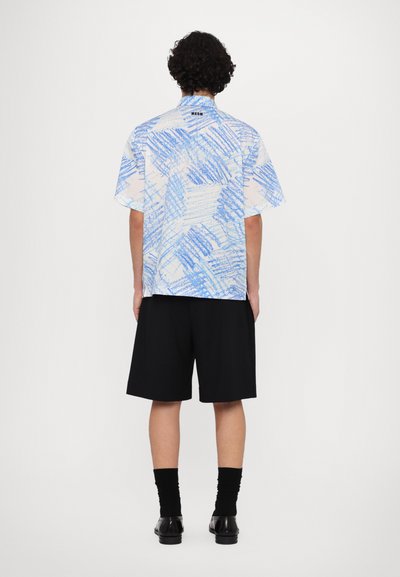 Camisa de manga corta en blanco con patrones abstractos azules, combinada con pantalones cortos negros, calcetines negros y zapatos negros; vista desde atrás.