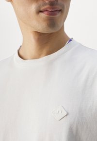 Biały bawełniany t-shirt z teksturowaną łatą w kształcie białego diamentu na lewej piersi. Gładka tkanina z klasycznym, okrągłym dekoltem.