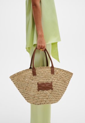 Γυναίκα ντυμένη με ανοιχτό πράσινο σύνολο κρατώντας μεγάλη υφασμένη τσάντα από άχυρο με καφέ δερμάτινες λαβές και πλακέτα.