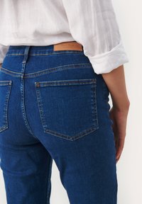 Tummansiniset farkut korkealla vyötäröllä, joissa on ruskea nahkainen logo-etiketti ja oranssit tikit takataskuissa. Sujuva denimgolfi.