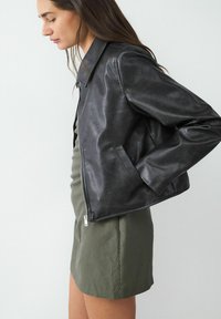 Giacca di pelle nera con una texture liscia, dotata di colletto, tasche laterali e una cerniera frontale. Indossata sopra un vestito a righe sottili color verde oliva.