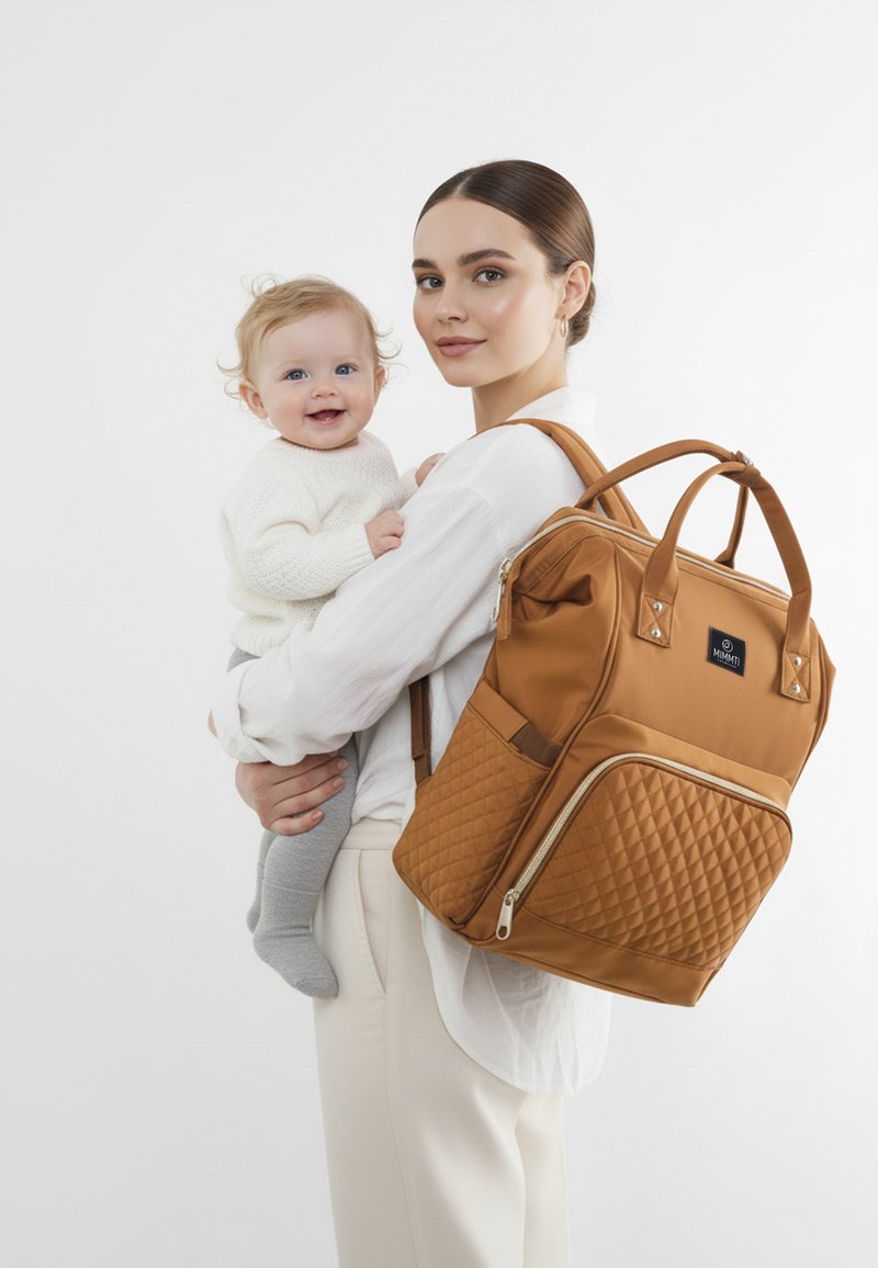 Brun quiltet pusletaske med dobbelthåndtag og en lynlåslomme, båret af en kvinde der holder en baby. Hvid baggrund; minimalistisk design.