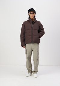 Doudoune marron avec un design matelassé, fermeture éclair argentée, portée sur un haut noir, associée à un pantalon cargo clair et des baskets blanches.