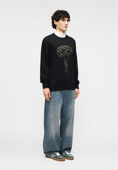 Μαύρο φούτερ με χρυστό ανθικό σχέδιο, σε συνδυασμό με φαρδιά μπλε τζιν και πράσινα αθλητικά παπούτσια με λευκά κορδόνια.