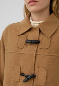Nahaufnahme eines kamelfarbenen Mantels mit schwarzen Knebelknöpfen, der über einem cremefarbenen Strickpullover getragen wird. Dabei sind der Kragen und der obere Brustbereich sichtbar.