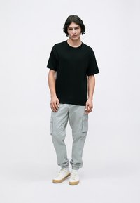 Črna kratka majica s kratkimi rokavi, lahke sivo cargo hlače z bočnimi žepi in bele superge z svetlo rjavimi podplati. Preprost in funkcionalen dizajn.