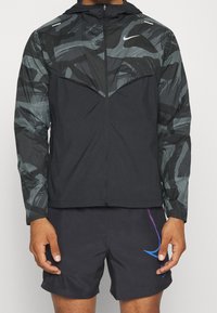 Nike windbreaker in zwart met een patroond bovenstuk in grijs, met een ritssluiting, capuchon en elastische manchetten; gedragen met donkere shorts.