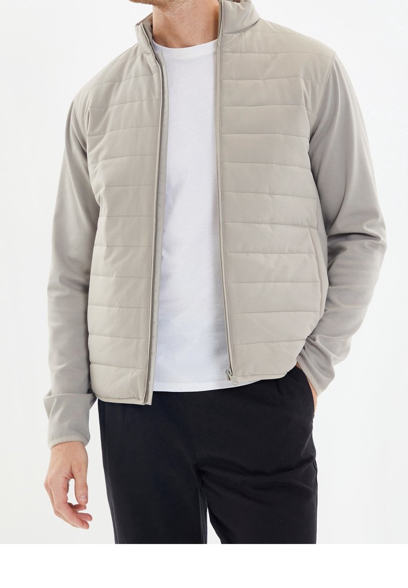 Homme portant une veste matelassée gris clair sur une chemise blanche avec un pantalon noir, debout avec une main dans la poche, sur fond blanc.