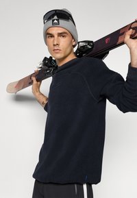 Giovane uomo con berretto grigio e maglione nero tiene gli sci sulla spalla, con occhiali da sole appoggiati sul berretto, guardando avanti.