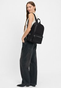 Zaino in pelle nera con design arrotondato, dotato di una tasca frontale con zip, spallacci regolabili e un manico superiore. Indossato con jeans scuri larghi.