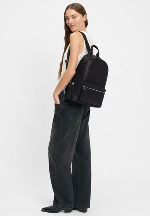 Zaino in pelle nera con design arrotondato, dotato di una tasca frontale con zip, spallacci regolabili e un manico superiore. Indossato con jeans scuri larghi.