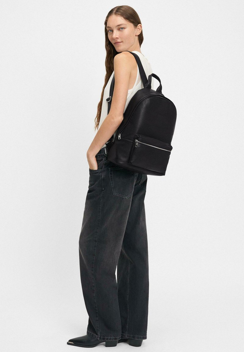 Zaino in pelle nera con design arrotondato, dotato di una tasca frontale con zip, spallacci regolabili e un manico superiore. Indossato con jeans scuri larghi.