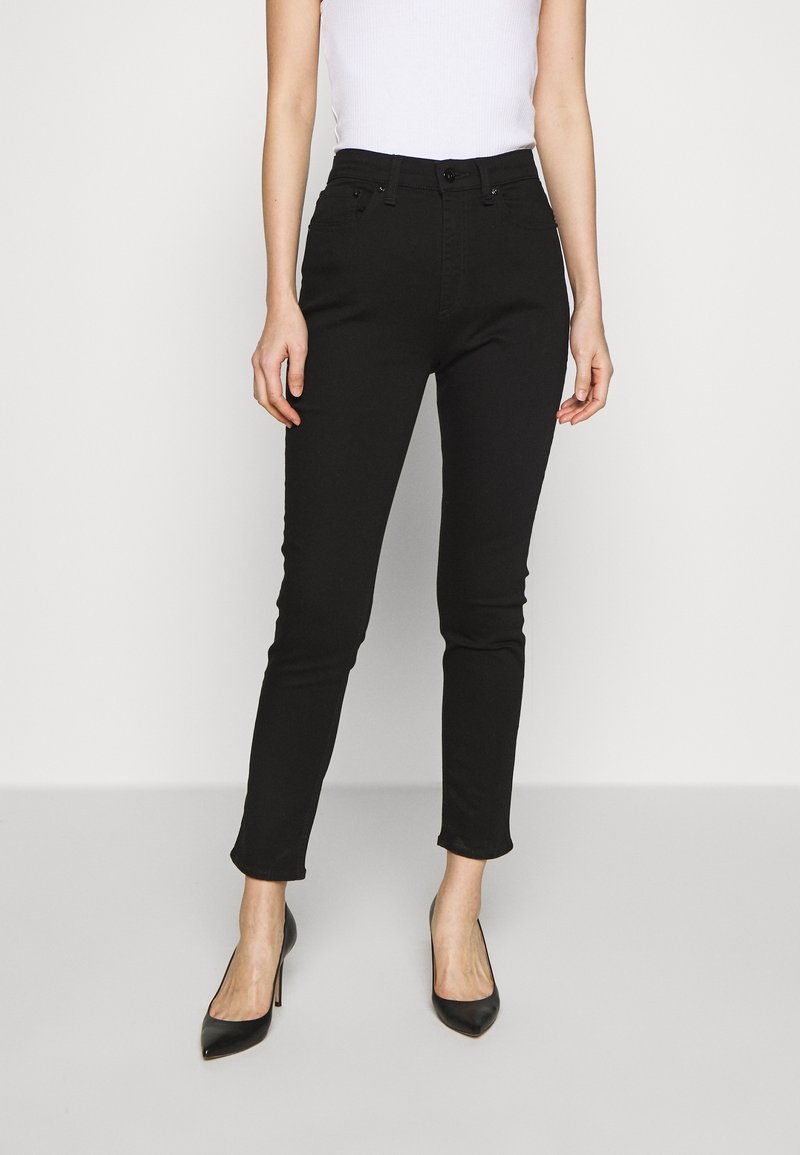 Mujer con jeans ajustados negros, tacones altos negros y una blusa blanca sin mangas, de pie contra un fondo blanco liso.