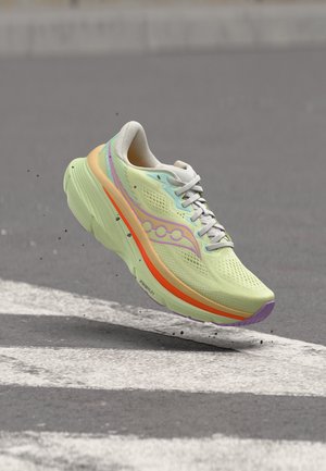 Zapatilla de correr verde claro con detalles en naranja y morado flotando sobre una carretera gris con rayas blancas y pequeños escombros.