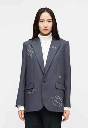 Γυναίκα με γκρι σακάκι στο οποίο έχουν κεντηθεί λευκά αστέρια, περιστέρια και κυματισμοί, φορώντας λευκό ζιβάγκο και σκοτεινό παντελόνι, στέκεται μπροστά σε λευκό φόντο.