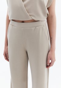 Beige, strukturovaný top s vázáním, doplněný o kalhoty s vysokým pasem ve stejném odstínu. Kalhoty mají hladký, přiléhavý střih s bočními kapsami.