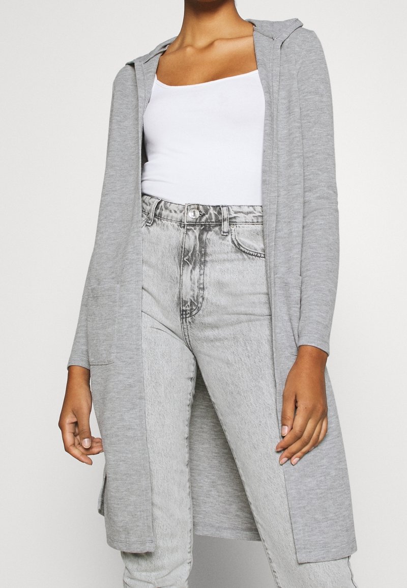 Szary, długi kardigan z kapturem, wykonany z miękkiego materiału. Posiada otwarty przód i dwie boczne kieszenie, zestawiony z jasnoszarymi dżinsami.