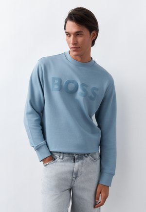 Άνδρας που φοράει ανοιχτό μπλε φούτερ με ανάγλυφα γράμματα "BOSS" και ανοιχτόχρωμο τζιν, στέκεται μπροστά σε λευκό, απλό φόντο.