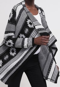 Černobílý oversize kardigan s geometrickými vzory, texturovanou látkou, otevřeným předním dílem a dlouhými rukávy. Výrazné třásně podél okrajů.