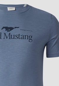 Niebieska koszulka z czarnym logo biegnącego mustanga i napisem "Mustangers zawsze wygrywają" oraz częściowo widocznym dużym napisem "Mustang".