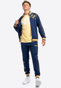 Ensemble de survêtement bleu marine avec des accents jaunes, comprenant une veste zippée et un pantalon assorti. La veste présente un design de logo ; le pantalon a un logo sur la jambe.