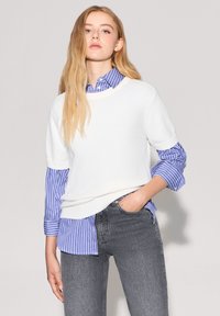 Biały sweter z dzianiny z krótkimi rękawami nałożony na niebieską i białą koszulę w paski, połączony ze szarymi dżinsami, eksponując teksturę i kontrast.