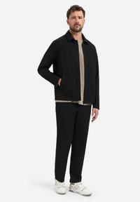 Mann in schwarzer Jacke und Hose, beiger Hemd und weißen Sneakers, der mit einer Hand in der Jackentasche vor weißem Hintergrund steht.