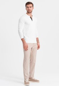 Mann steht da und trägt ein weißes Langarm-Henley-Shirt, beige Hosen, beige Sneakers und ein Perlenarmband am linken Handgelenk.