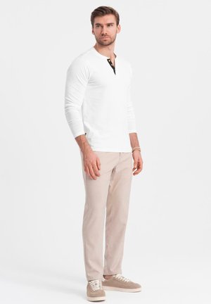 Hombre de pie vistiendo una camisa henley blanca de manga larga, pantalones beige, zapatillas beige y una pulsera de cuentas en la muñeca izquierda.