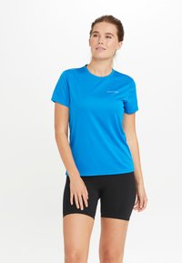 Sininen urheilu t-paita, joka on valmistettu kevyestä kankaasta ja jossa on lyhyet hihat. Ominaisuuksiin kuuluu pyöreä kaula-aukko ja huomaamaton logo rinnassa. Mustat shortsit.