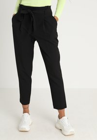 Musta räätälöity housut, jotka imevät vyötärönauhan, pleidatut etuosa ja kapenevat nilkat, yhdistettynä valkoisiin paksupohjaisiin lenkkareihin. Sujuva kankaan rakenne.