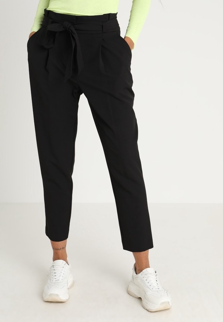 Musta räätälöity housut, jotka imevät vyötärönauhan, pleidatut etuosa ja kapenevat nilkat, yhdistettynä valkoisiin paksupohjaisiin lenkkareihin. Sujuva kankaan rakenne.